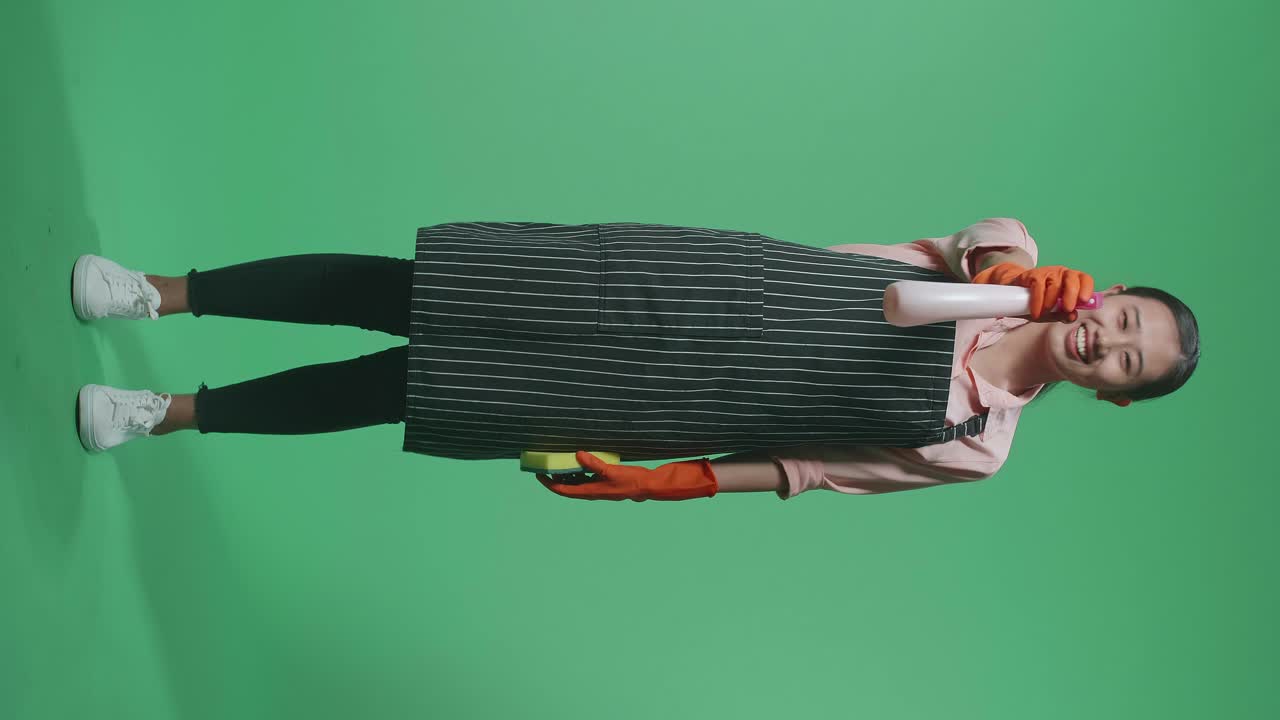 미소 짓는 아시아 여성 집주인이 주방 스폰지를 들고 녹색 스크린 스튜디오에 서있는 동안 스프레이를 사용합니다.