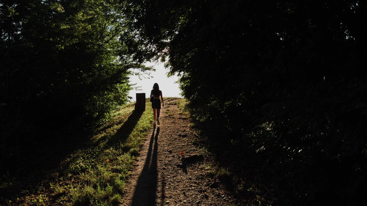 Una giovane donna cammina lungo uno stretto sentiero nel bosco in estate, muovendosi verso la luce solare intensa tra alberi scuri, foglie verdi e lunghe ombre.