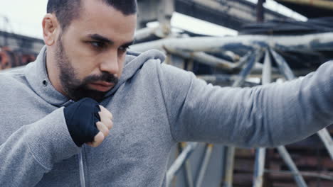 close-up view of caucasian man in sportswear hitting a punching bag outdoors an abandoned factory on a cloudy morning