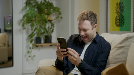 man relaxing on the sofa in his home, laughing at some funny video viewed on his cell phone