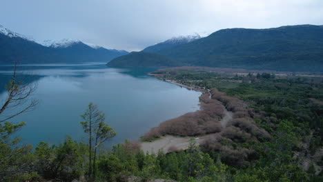 Panoramic-view-of-the-lake,-forest-and-snowy-mountains-in-cloudy-day-panning-right