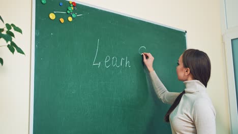 young english teacher is writing on the board. beautiful woman in sweater writes the english words with a chalk. the words learn english is written on the blackboard by teacher.