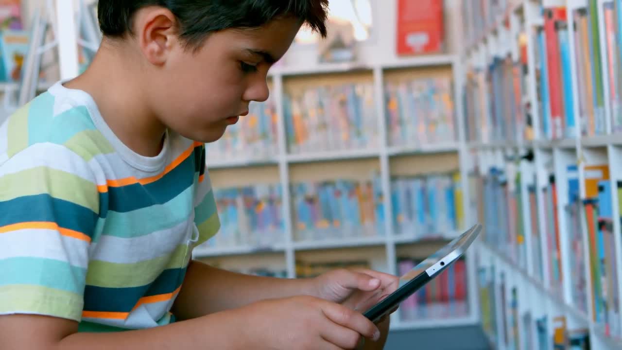 escolar caucásico sentado en el sofá y usando tableta digital en la biblioteca de la escuela 4k