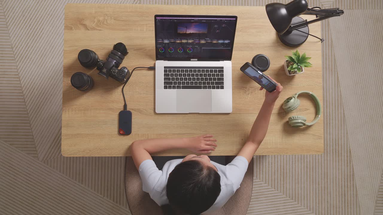 vista superior de una mujer clasificando el color agitando la mano y haciendo una llamada de video en el teléfono inteligente mientras está sentada en el espacio de trabajo usando una computadora portátil al lado de la cámara editando el video en casa