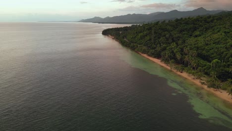 Toma-Aérea-De-Un-Dron-De-La-Tranquila-Costa-Del-Océano-Junto-A-Una-Pintoresca-Isla-Tropical-Con-Exuberantes-Palmeras-Al-Atardecer