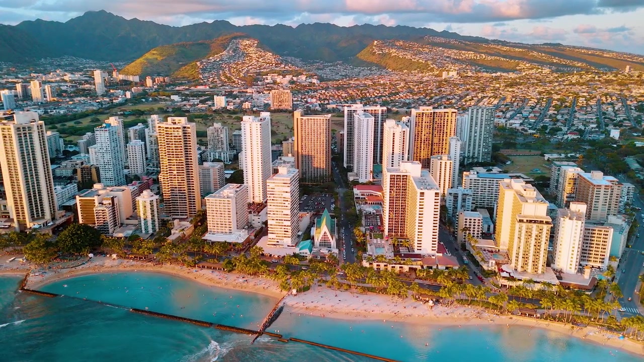Luce dell'ora d'oro sugli hotel della spiaggia di Waikiki, Hawaii. Vista elevata di una fila di resort fronte mare illuminata, costa sabbiosa, acqua turchese e viale alberato di palme, con catena montuosa sullo sfondo.