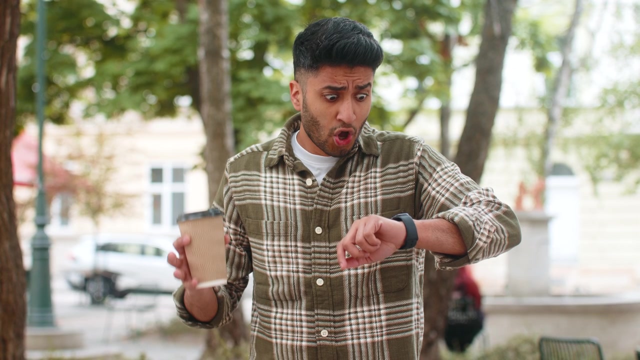 Indian man holding coffee walking in city street checking wristwatch feeling annoyed before meeting