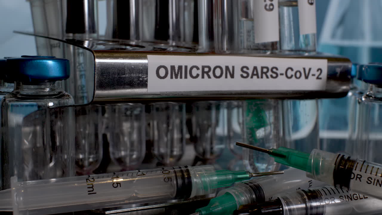 A close look on a metal tube rack labeled OMICRON SARS-CoV-2. The glass test tubes named B.1.1.529 are taken one by one from the rack. Stacked syringes lie in front of a rack. The background is blurry