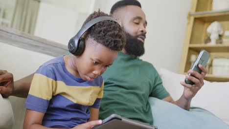 Feliz-Padre-E-Hijo-Afroamericanos-Sentados-En-El-Sofá,-Usando-Teléfonos-Inteligentes-Y-Tabletas,-En-Cámara-Lenta