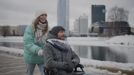 Glückliche-Kaukasische-Frau,-Die-Ihren-Behinderten-Freund-Im-Rollstuhl-Für-Einen-Spaziergang-In-Der-Stadt-Nimmt-Und-Miteinander-Redet