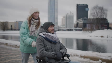 happy caucasian woman taking her disabled friend in wheelchair for a walk in the city and talking together