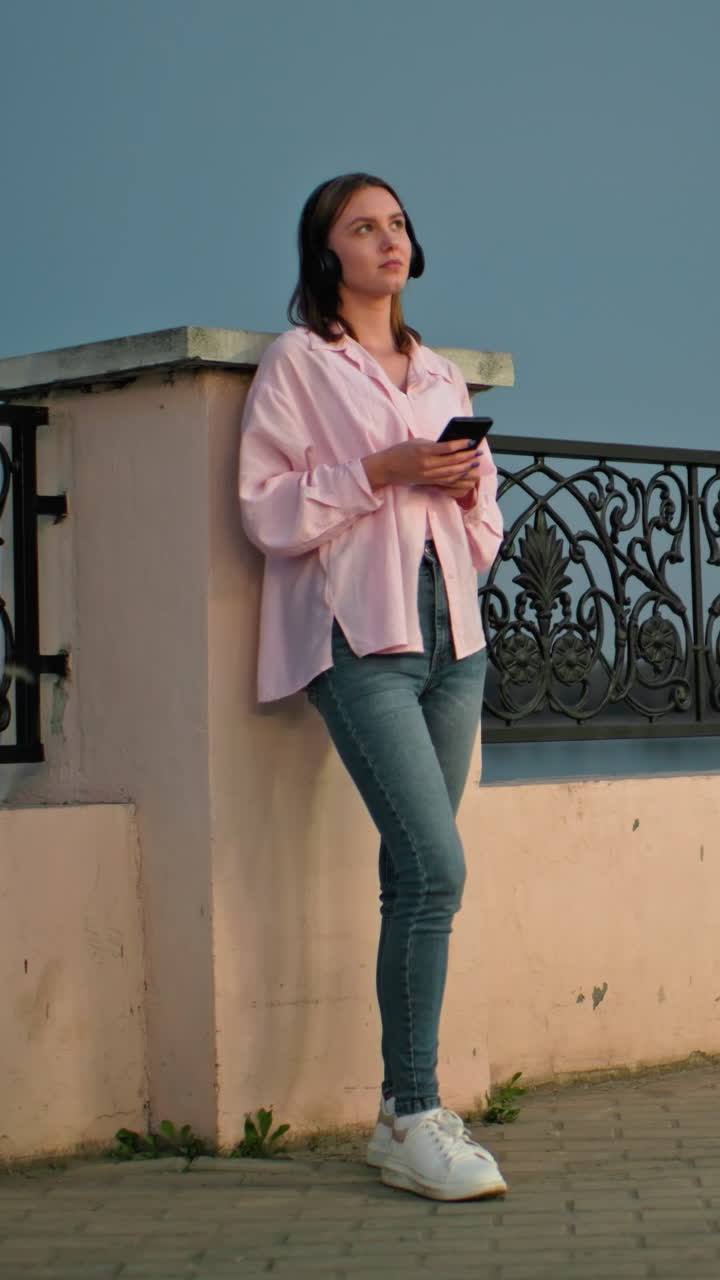 Woman standing outdoors at dusk, leaning against decorative iron fence, wearing headphones and holding phone, soft evening lighting with moon visible in background