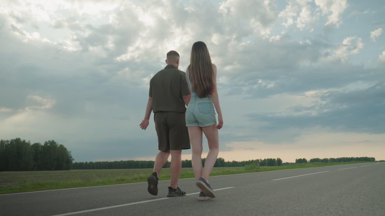 rear view of couple walking hand in hand along empty country road under dramatic cloudscape at sunset, long hair swaying in casual summer attire, evoking freedom