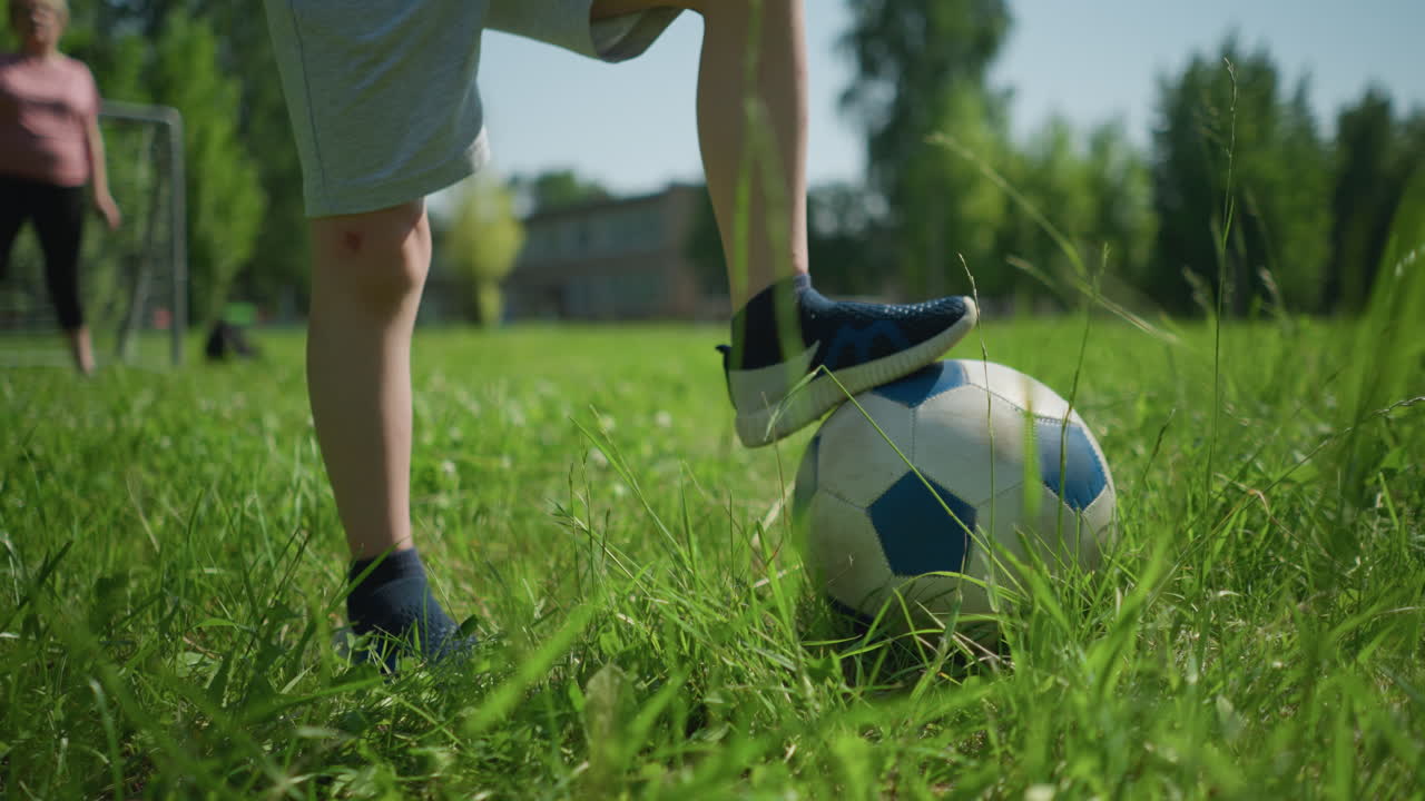 vista cercana de la pierna de un niño pequeño colocando un pie en una pelota de fútbol en un campo cubierto de hierba, con una vista borrosa de una mujer de pie frente a un poste de la portería