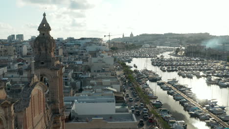 Small-village-in-Malta-revealing-behind-two-Church-Bell-towers-with-boats-in-port-on-Sunny-day,-Aerial-View