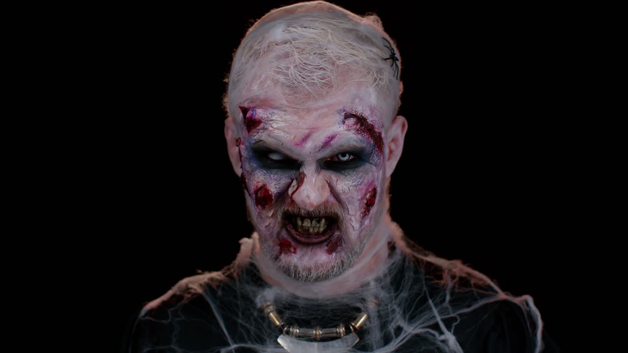 Zombie man with makeup with fake wounds scars and white contact lenses looking at camera, screaming
