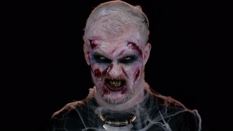hombre zombi con maquillaje con heridas falsas cicatrices y lentes de contacto blancas mirando a la cámara, gritando