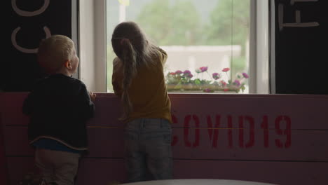 little girl waves hand and has fun with brother near window