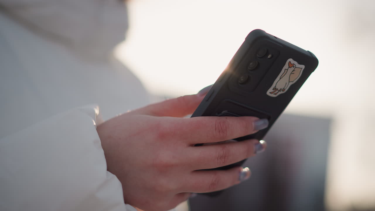Smart phone with black case held by manicured hand scrolling through apps as young creative strolls through snowy park wearing winter coat headphones and tinted goggles under glowing sunlight