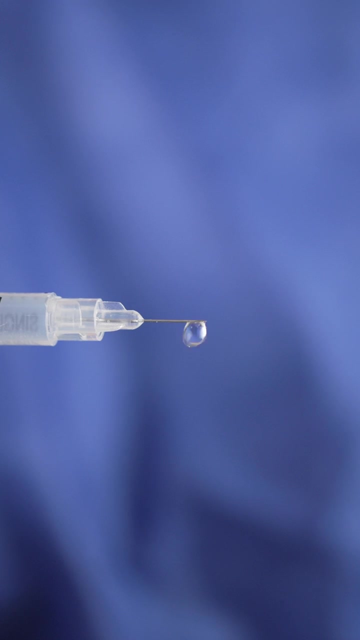 Vista macro ravvicinata di una sottile punta di ago ipodermico che trattiene una gocciolina di liquido trasparente su uno sfondo di tessuto blu morbido, utile per temi di vaccinazione, sicurezza delle iniezioni o assistenza sanitaria.