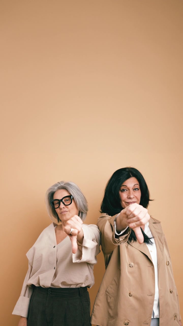 Two Mature Women Giving Thumbs Down Gesture Against Beige Background