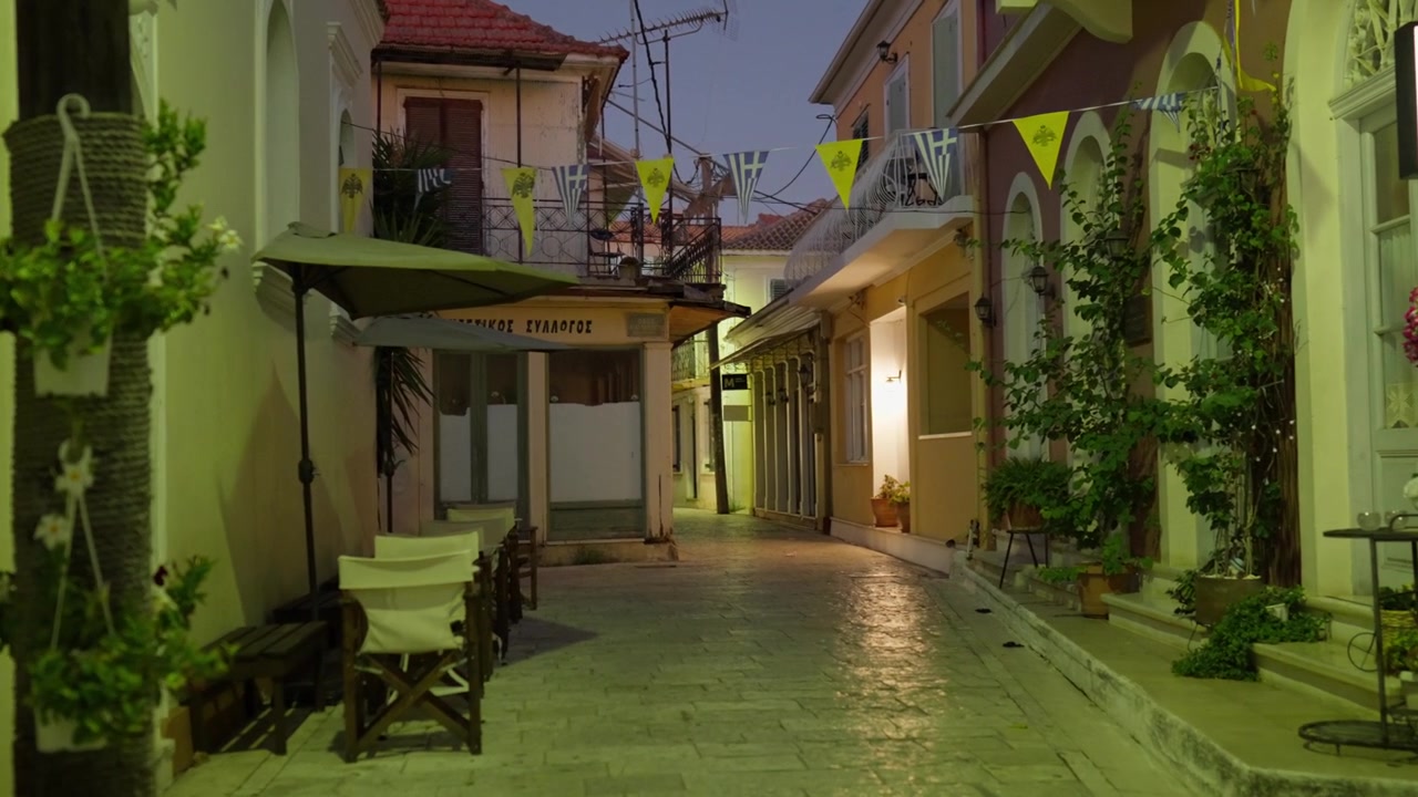 Narrow paved alleyway with yellow buildings, decorative flags, and warm street lights at twilight.