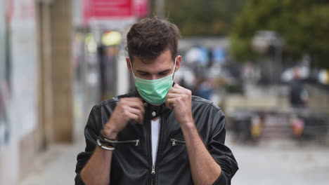 a young man with a cool hairstyle wearing a black jacket and a white t-shirt, putting on a green protective covid-19 facemask on a busy street, looking straight into the camera, static close up 4k