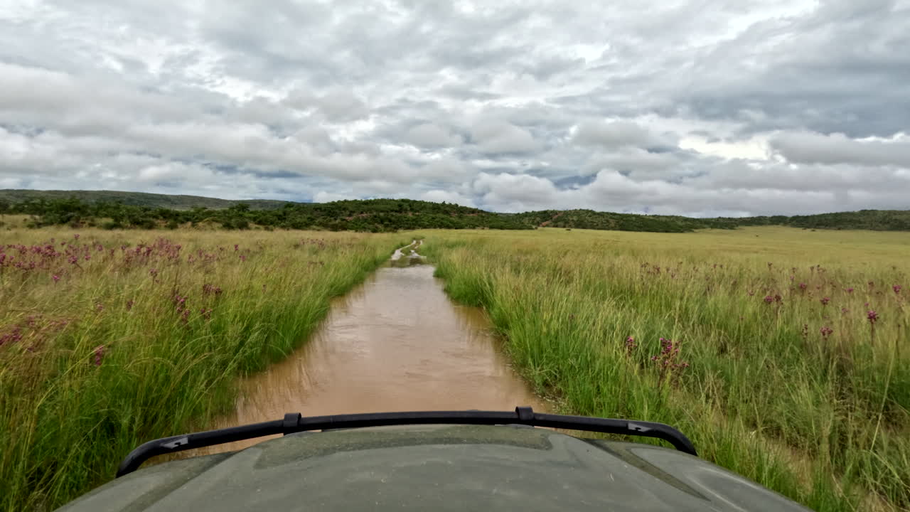 Flooded dirt road in Africa with water splashing, tall grass, and stormy clouds ahead
