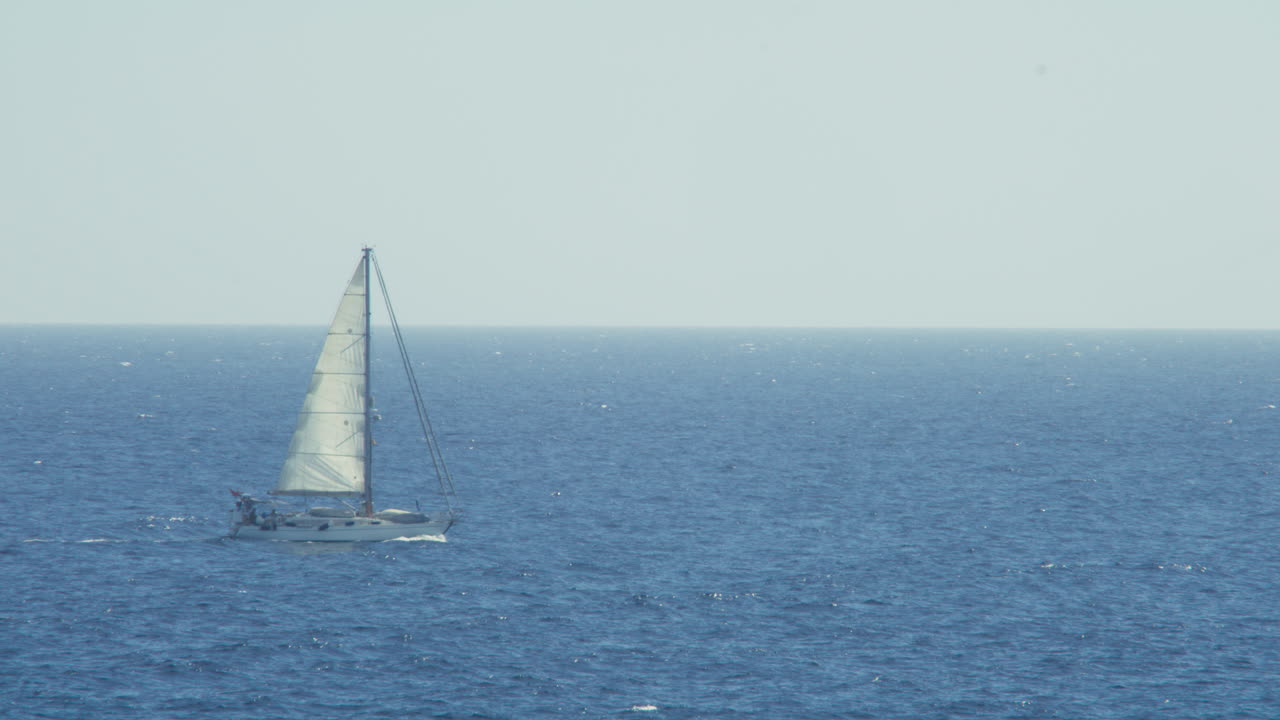 Wide static shot showing a sailboat gliding calmly across the deep blue sea from left to righ. Evoking freedom, movement, and peaceful isolation