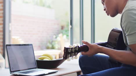 Un-Hombre-Afroamericano-Toca-La-Guitarra-Y-Canta,-Usando-Una-Computadora-Portátil-En-Casa