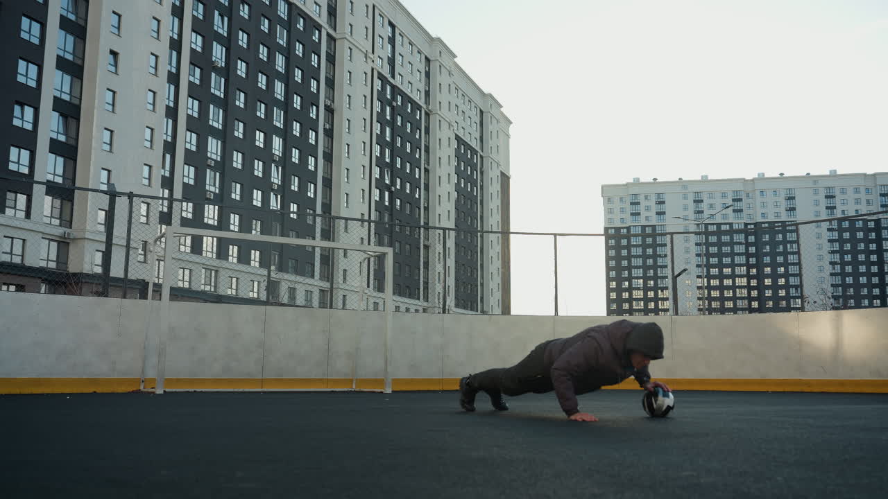 atleta realizando flexiones dinámicas alternando la colocación de la mano en una pelota de fútbol demostrando fuerza, coordinación y agilidad contra un telón de fondo de postes de gol y edificios residenciales