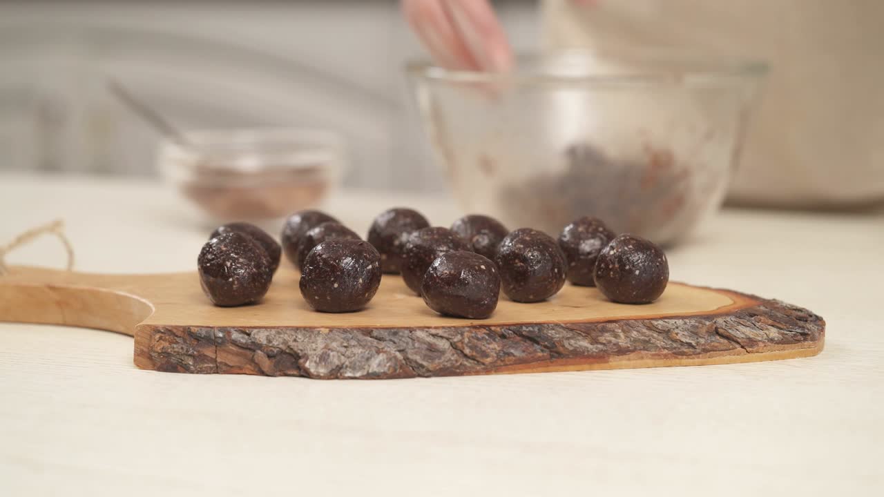 Bottom top shot of homemade round chocolate sweets on the wooden cooking board in the foreground. Woman's hand putting a candy onto the board. Close-up demonstration video