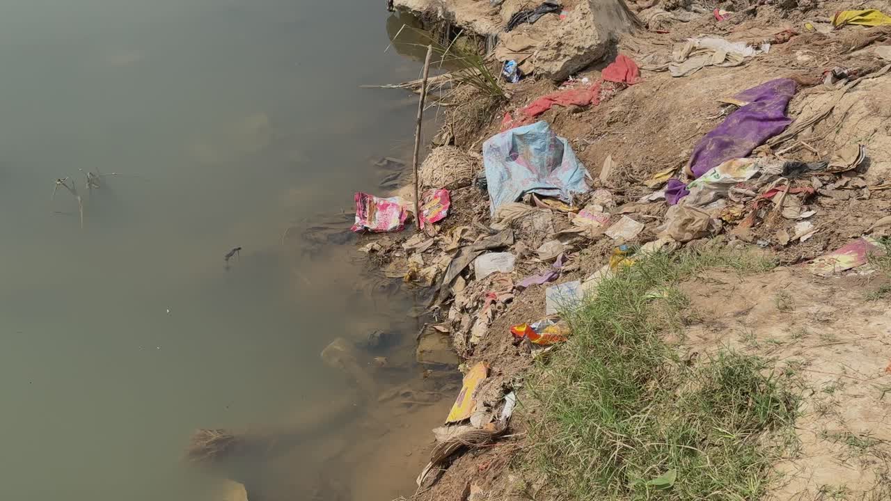 A static view of a polluted riverbank covered with discarded fabrics, plastic waste, and debris, contrasting against the murky water and sparse patches of grass