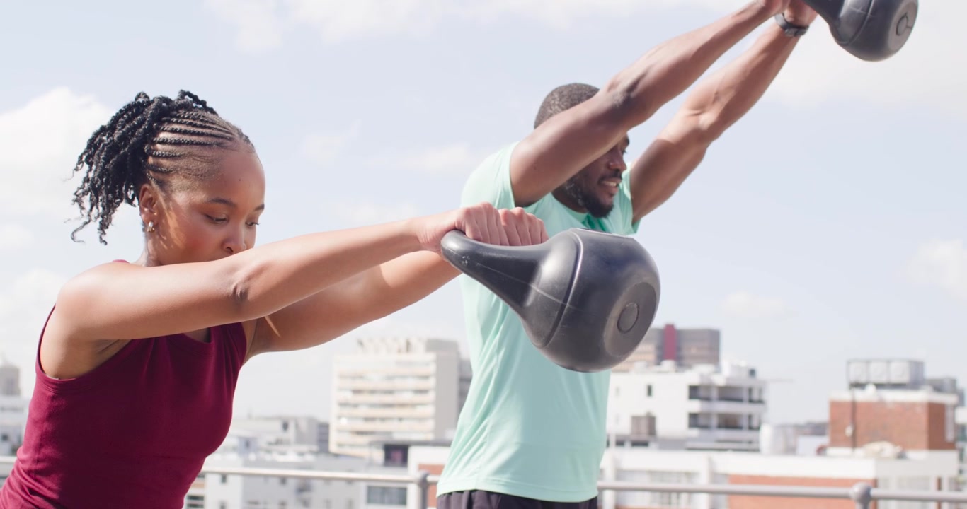 African American workout partners in gymwear reaching for 15kg kettlebells on roof swinging, toning