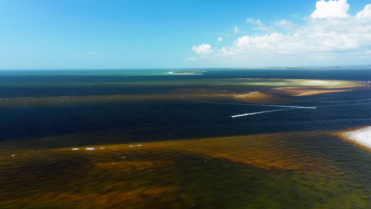 Offshore sandbars scatter across emerald-green and deep blue waters near a sandy peninsula, forming striking natural patterns where shallow and deep seas converge
