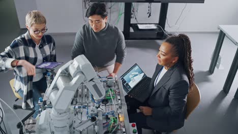 black teacher explaining information to students gathering around table with robot limb. people holding devices with apps and discussing it at university. education concept. high angle view