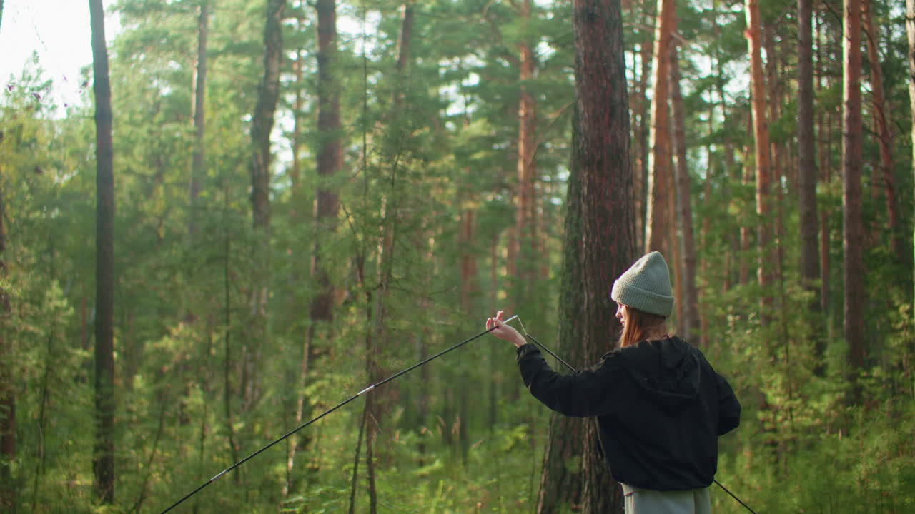 Lady in hoodie and beanie fixing tent pole with forest background as another person holds pole from side, showcasing teamwork and outdoor camping preparation in soft natural light