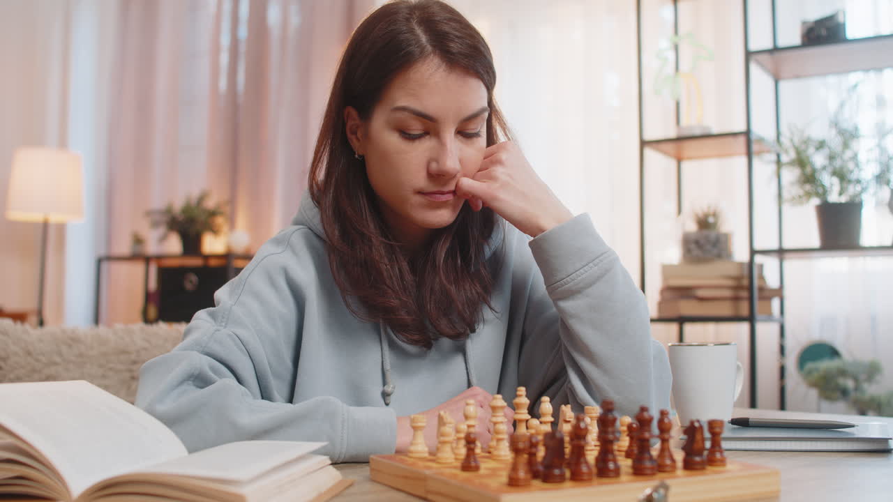 joven jugando al ajedrez sola arreglando piezas en la mesa en el sofá con el libro acostado cerca en casa