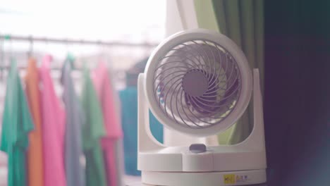 mini portable electric fan in white and purple color rotating inside a room in the house with clothes hanging in the blurry background - slow motion