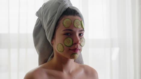 portrait of woman with cucumber slices placed on face and hair wrapped in towel, relaxing during rejuvenating skincare and self-care moment, calm expression focused on wellness and natural beauty