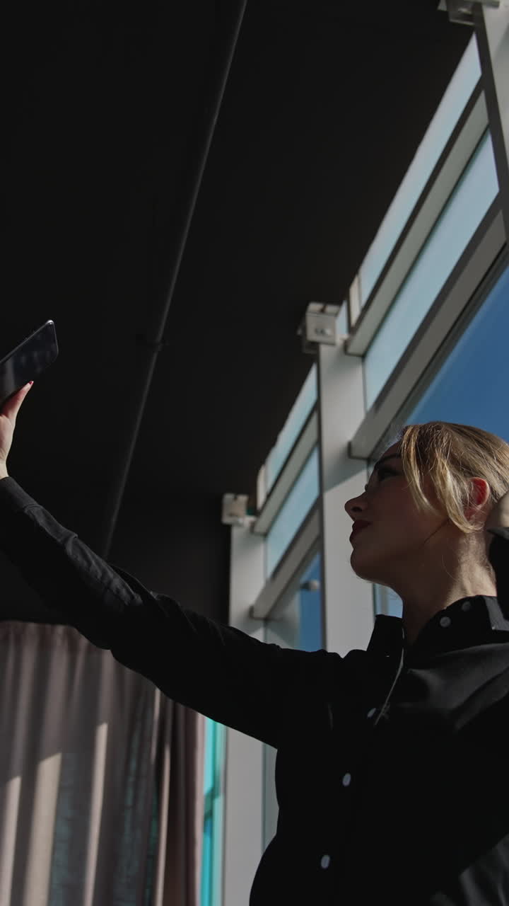 Attractive woman holding a smartphone in front of herself. Lady is posing in front of phone camera in a sunny office. View from below. Vertical video