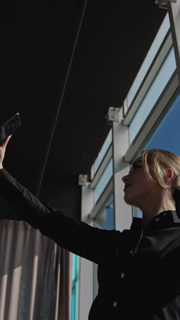 attractive woman holding a smartphone in front of herself. lady is posing in front of phone camera in a sunny office. view from below. vertical video