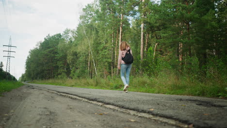 rear view of tourist walking along empty rural road surrounded by tall pine trees, carrying backpack and exploring forest area under soft daylight