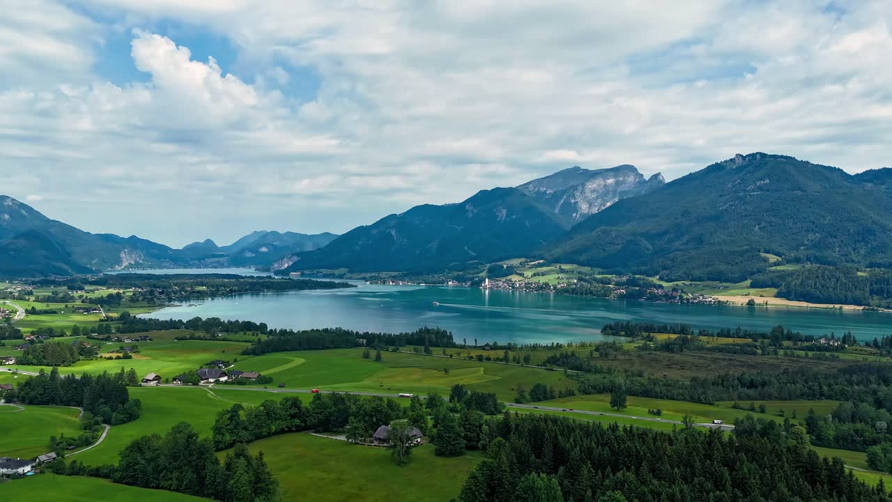 Fast-moving hyperlapse glides toward Lake Wolfgangsee under shifting midday clouds; boats skim turquoise water as sunbeams break through over the Salzburg Alps