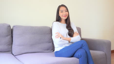 portrait of satisfied asian woman with crossed arms sitting on sofa and looking at camera with smile, full frame slow motion