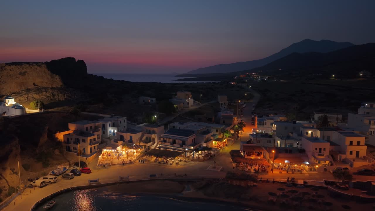 Aerial drone pulls back over Finiki village in Karpathos, showcasing the glowing lights, colorful sky, and serene Aegean twilight atmosphere