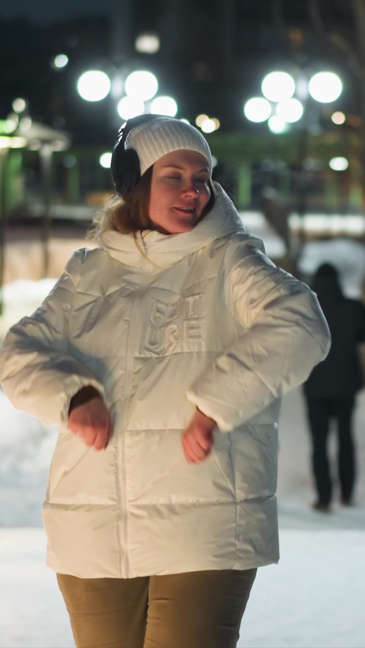 Woman wearing white puffer jacket with FUTURE text and black headphones dances outdoors at night in urban setting surrounded by bright streetlights. Woman moves energetically through scene