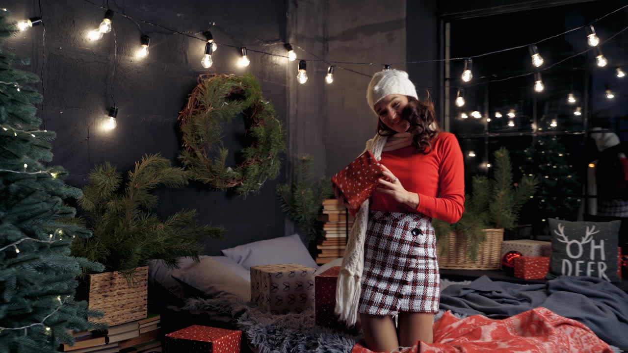 Happy girl on bed tossing a gift box. Beautiful young woman in knitted cap holding Christmas presents and having fun at home. New Year time.