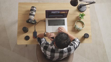 top view of a male editor talking on smartphone while sitting in the workspace using a laptop next to the camera editing the video at home