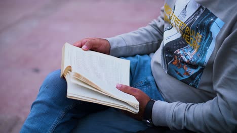 Toma-De-Primer-Plano-De-Un-Hombre-No-Identificable-Del-Sur-De-Asia-Leyendo-Un-Libro-Sentado-Afuera---Ratón-De-Biblioteca-Masculino-Disfrutando-De-La-Naturaleza-Mientras-Lee,-Joven-Del-Sur-De-Asia-Absorto-En-La-Lectura-Al-Aire-Libre